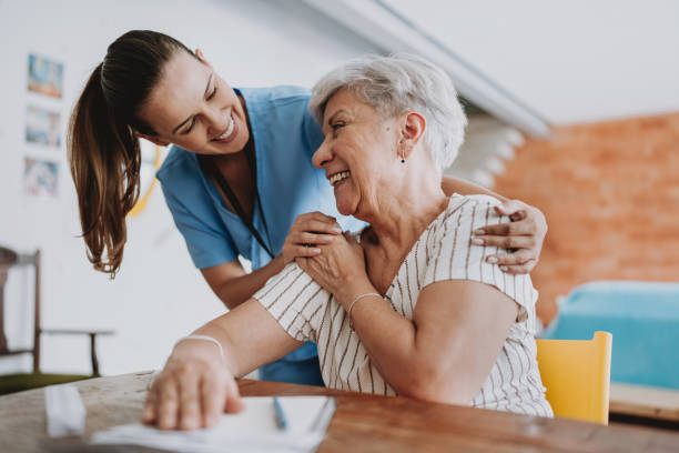 A nurse in scrubs resting a hand on the shoulder of a smiling older woman seated at a table, the two of them laughing together.